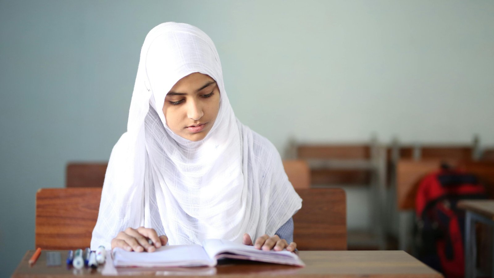 Young girl in hijab reading at a desk in a classroom. Focused and studious.